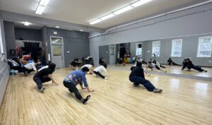 Participants stretch low to the floor in a mirrored dance studio, hands and feet spread wide.