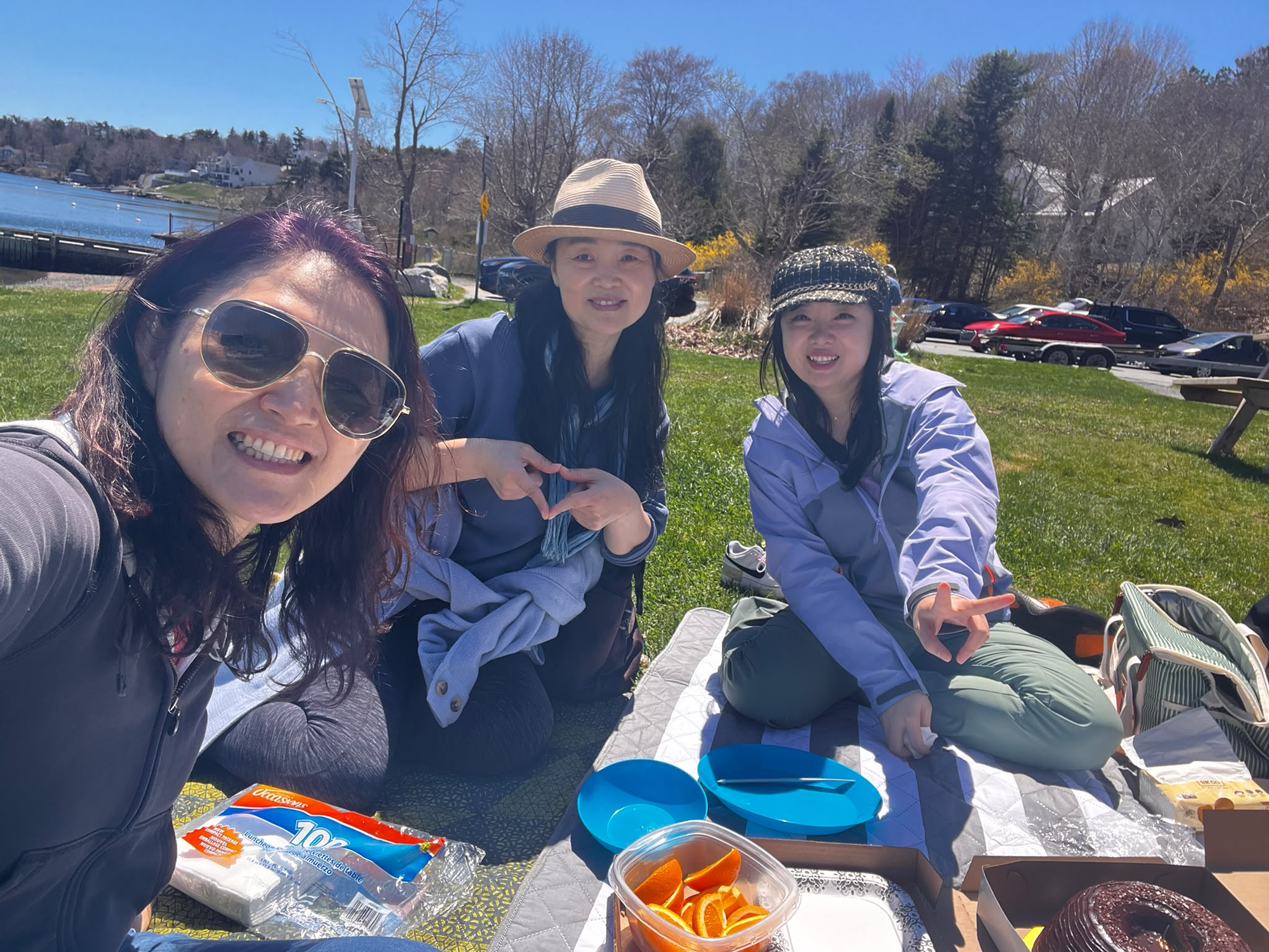 Three women pose for a selfie on a picnic blanket by the water during the Hiking & Picnic in the Park.