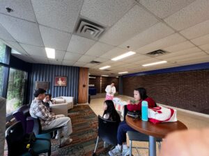 Ellie speaks near the table indoors while participants sit and listen nearby.