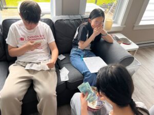 Participants sit on a couch counting cash and coins from a tin, with receipts nearby.