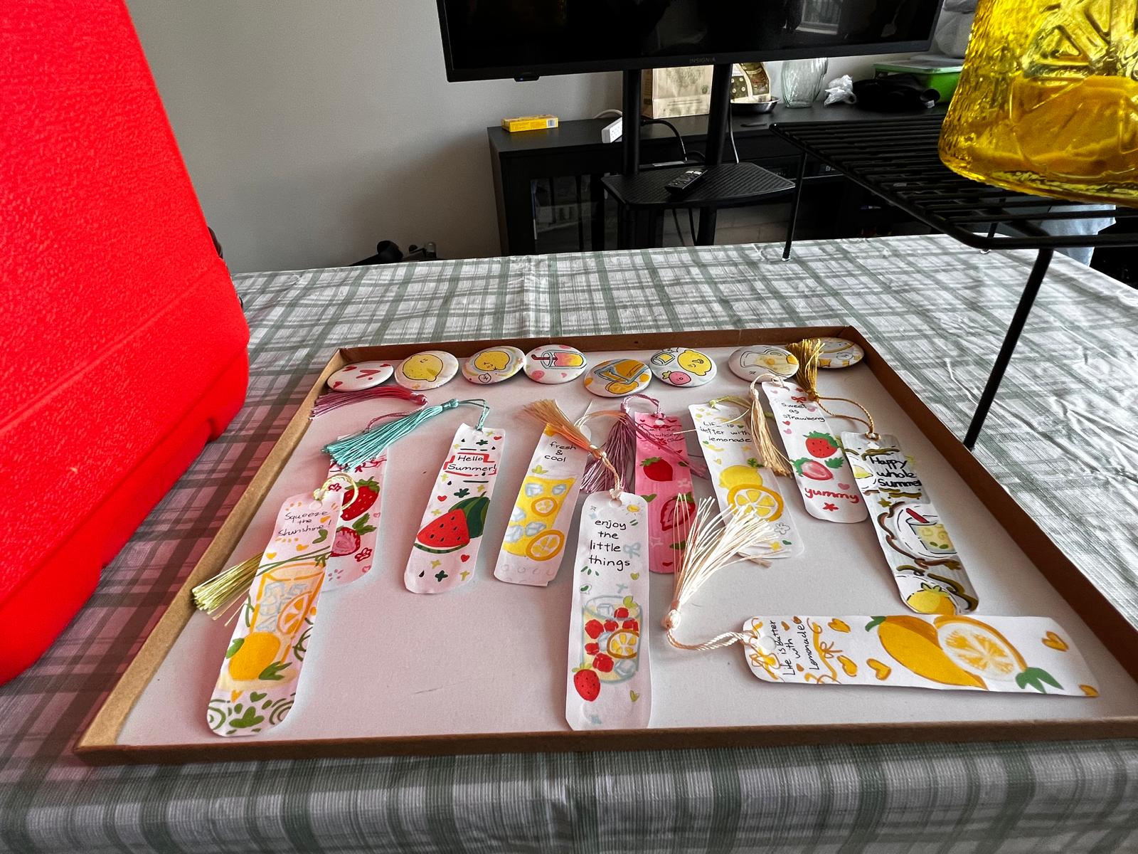 A tray displays handmade bookmarks and buttons with summer and lemonade-themed designs.