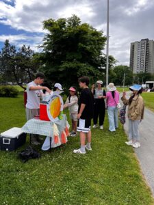 A teen serves drinks at an outdoor lemonade stand as customers line up on the grass.