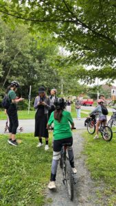 Group with bicycles and helmets gathers on a grassy trailside path under trees.