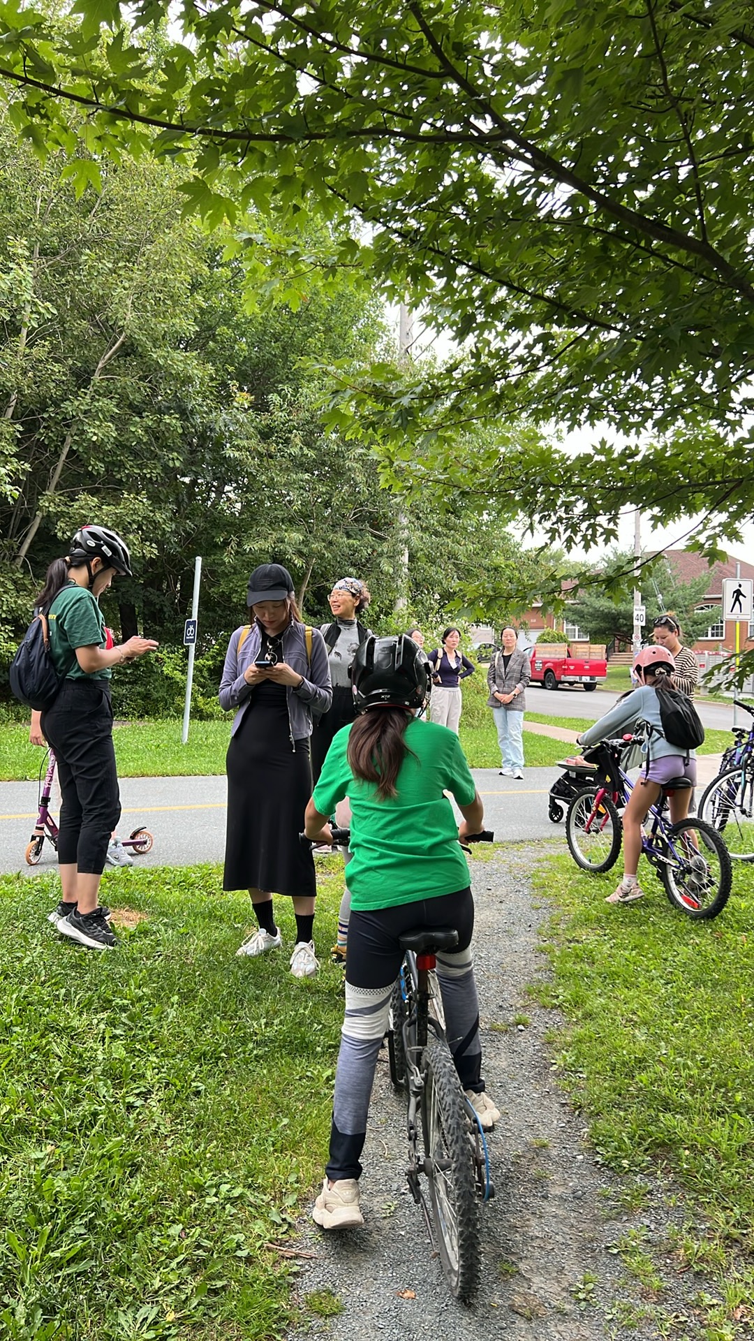 Group with bicycles and helmets gathers on a grassy trailside path under trees.
